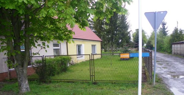 Zdjęcie przedstawiające teren będący przedmiotem najmu, ogrodzony płotem teren i przylegający do niego budynek oraz zieleń.